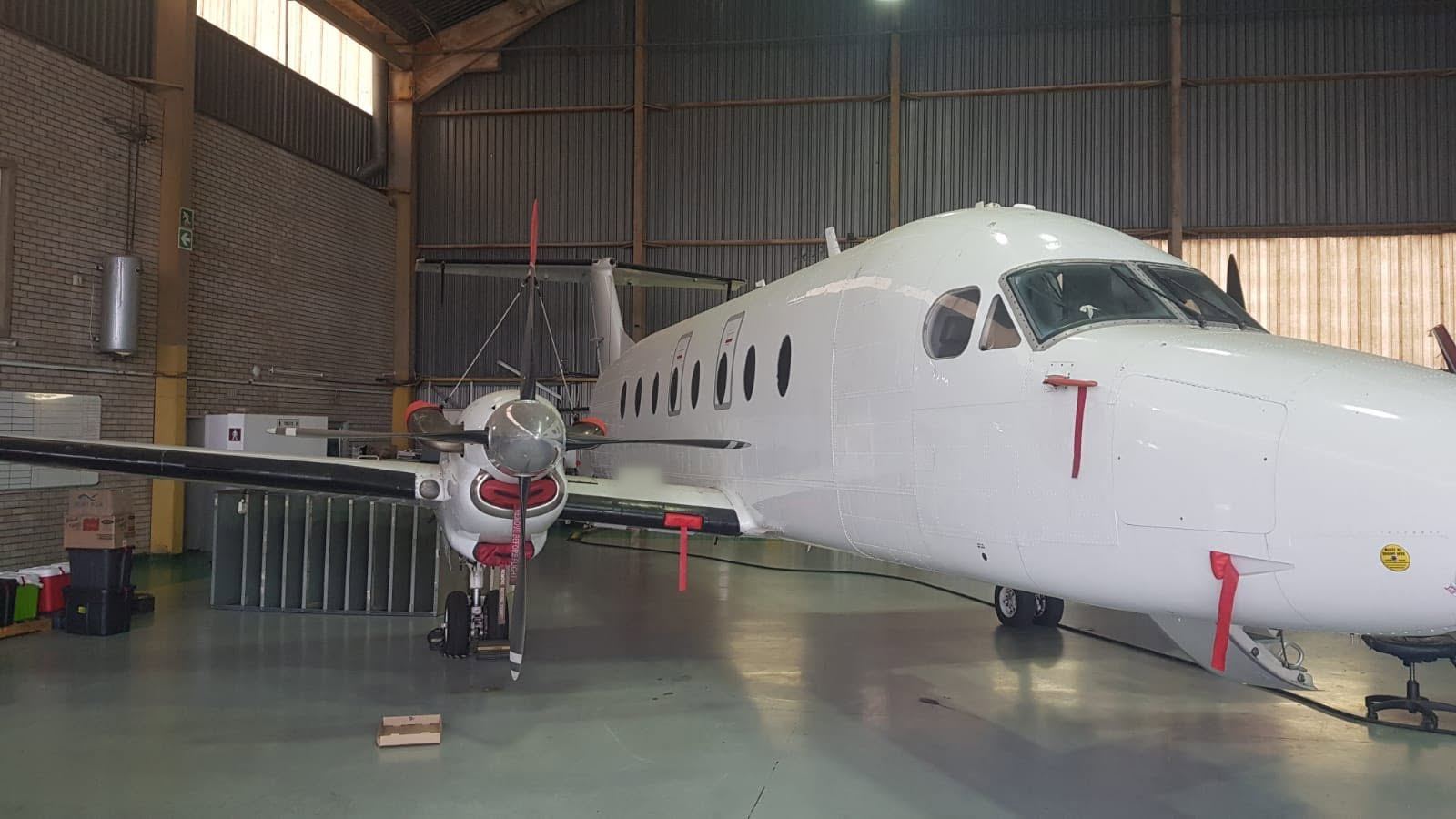 A white Beechcraft 1900D in a hangar, with red remove-before-flight streamers on the pitot covers and engine inlets.