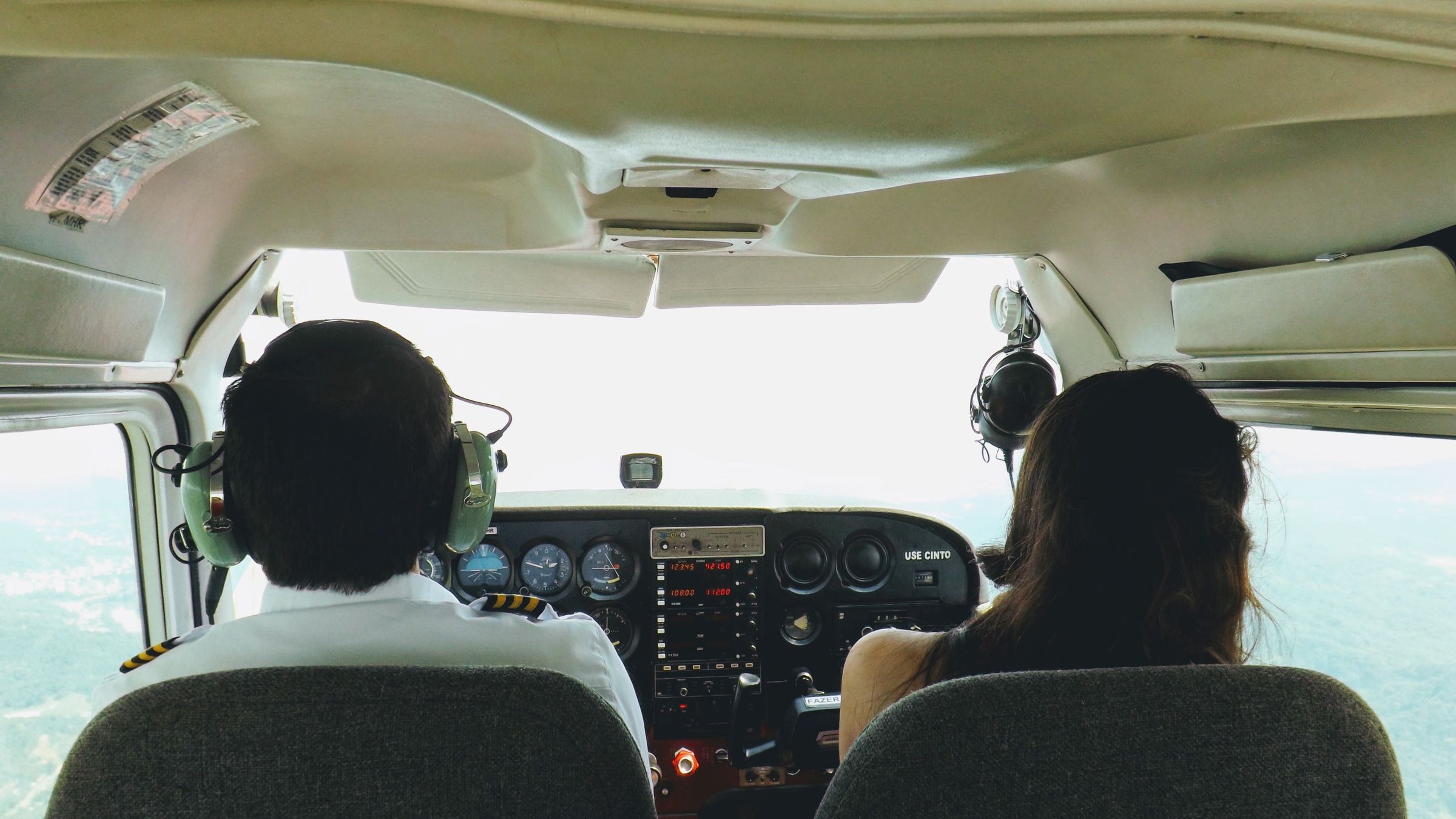 Two people seated side by side in the cockpit of a light aircraft, seen from behind.
